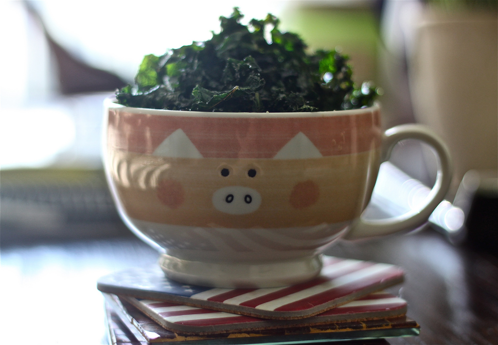 This image shows a close-up of a decorative mug standing on a stack of coasters on a counter top. The mug has a cartoon picture of a pig's face on it, and the mug is filled with lots of lettuce which is visible poking out of the top of the mug.
